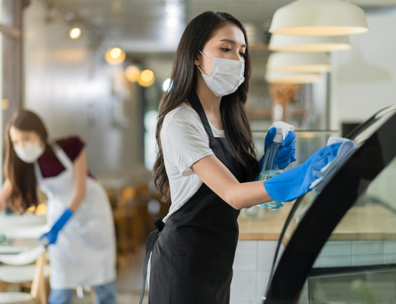 attractive positive young Asian woman staff in uniform apron spraying detergent or alcohol on counter product cabinet  wiping it with soft cloth when disinfecting cafe in ther morning safe and clean